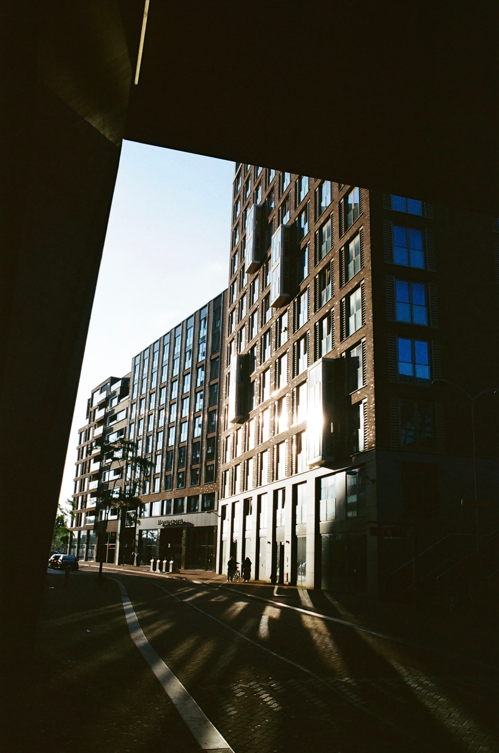 A city street with modern buildings, featuring a mix of glass and brick facades, under a clear blue sky. Shadows are cast on the ground, suggesting early morning or late afternoon sunlight
