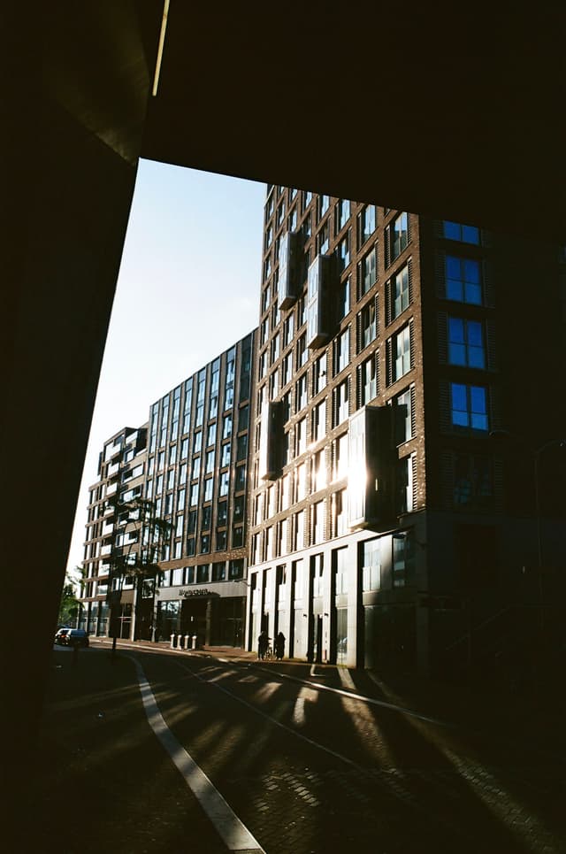 A city street with modern buildings, featuring a mix of glass and brick facades, under a clear blue sky. Shadows are cast on the ground, suggesting early morning or late afternoon sunlight