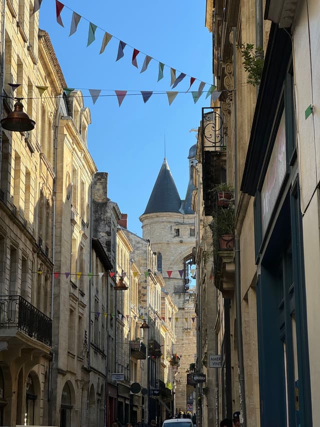A narrow European street lined with historic buildings, featuring a tower with a conical roof in the background and colorful pennant flags strung across the street