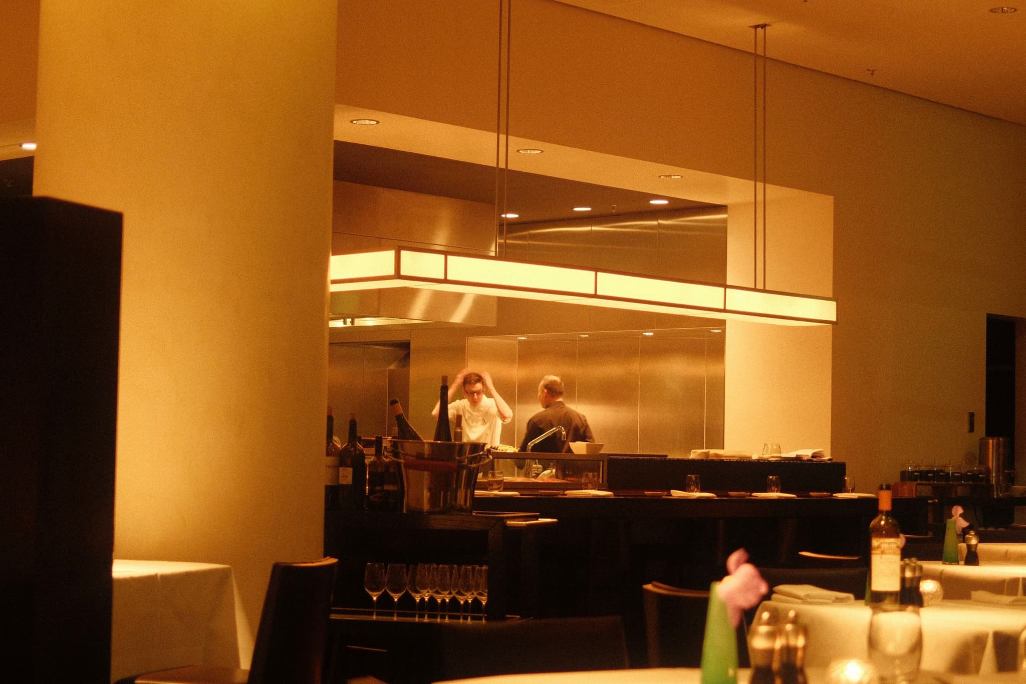 A restaurant kitchen with chefs working, viewed from the dining area. The lighting is warm, and tables are set with bottles and condiments