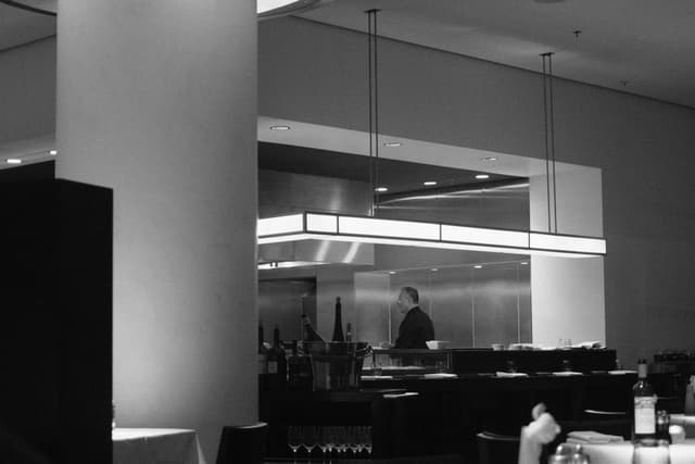 A black and white photograph of a modern restaurant interior with a visible kitchen area, featuring a chef working behind a counter
