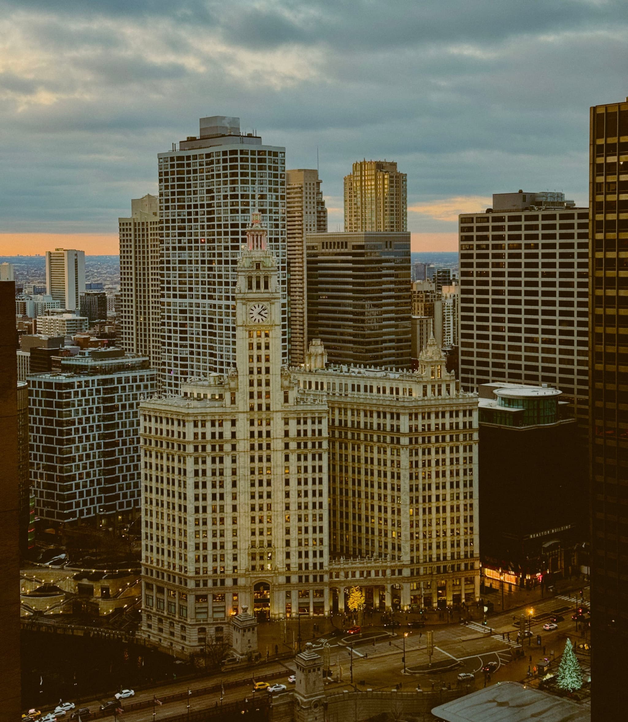A cityscape featuring tall buildings under a cloudy sky, with a prominent clock tower in the foreground and a hint of sunset on the horizon