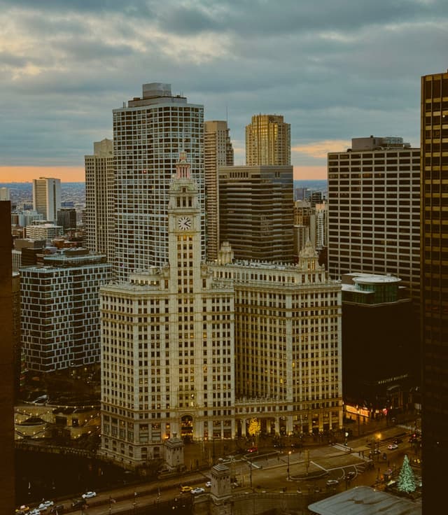 A cityscape featuring tall buildings under a cloudy sky, with a prominent clock tower in the foreground and a hint of sunset on the horizon