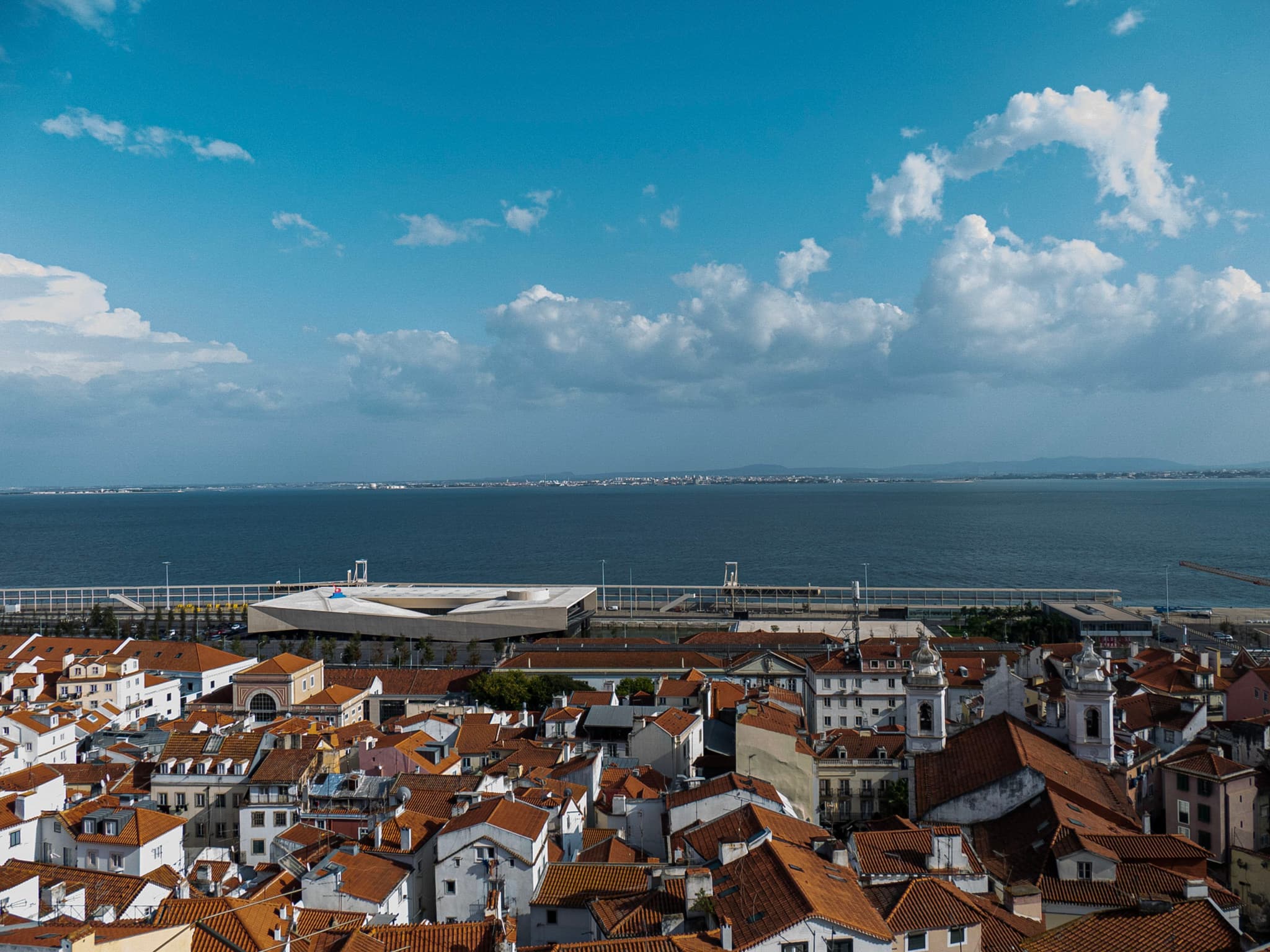 A coastal cityscape with red-roofed buildings in the foreground and a large body of water in the background under a partly cloudy sky
