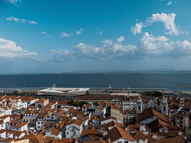A coastal cityscape with red-roofed buildings in the foreground and a large body of water in the background under a partly cloudy sky