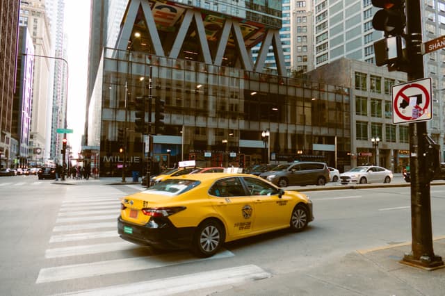 A yellow taxi driving through a city intersection with tall buildings in the background and a no U-turn sign visible