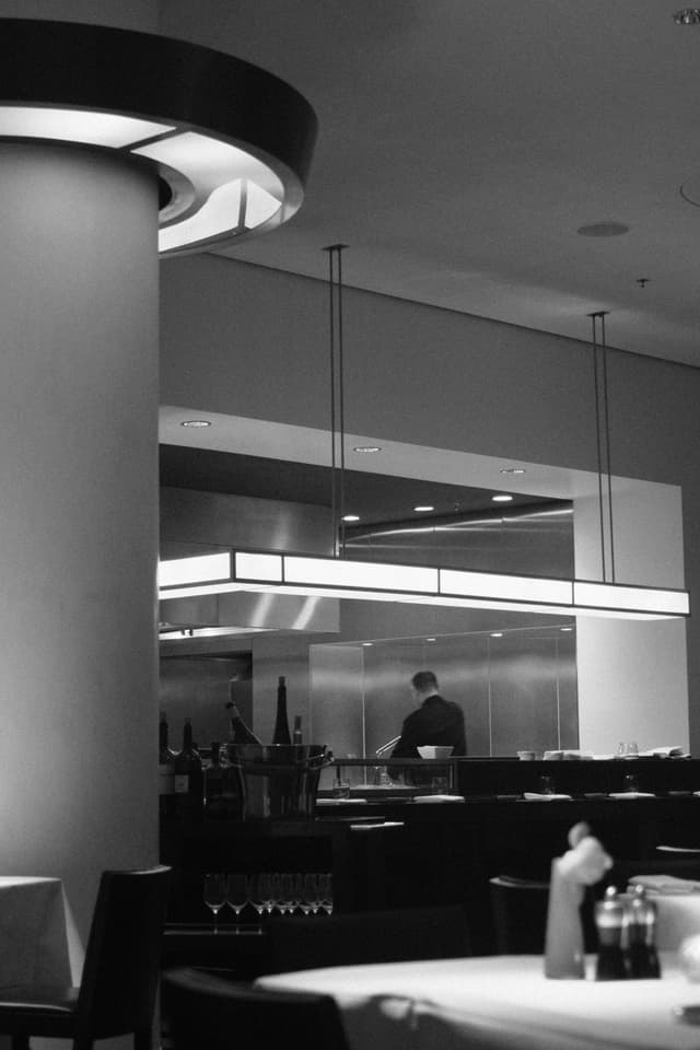 A black and white photo of a modern restaurant interior with a visible kitchen area, where a person is working. Tables are set with tablecloths and condiments