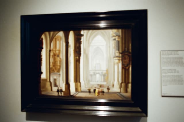 A framed painting of a grand, ornate interior with tall arches and columns, displayed on a wall next to a descriptive plaque