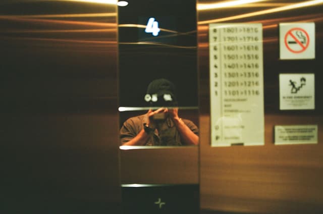 An elevator interior with a reflective panel showing a person taking a photo, floor number 4 displayed, and various signs on the wall