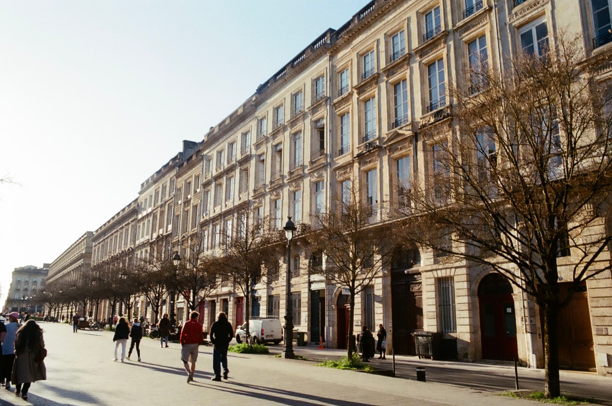 A sunlit street with people walking along a row of elegant, multi-story buildings and leafless trees lining the sidewalk