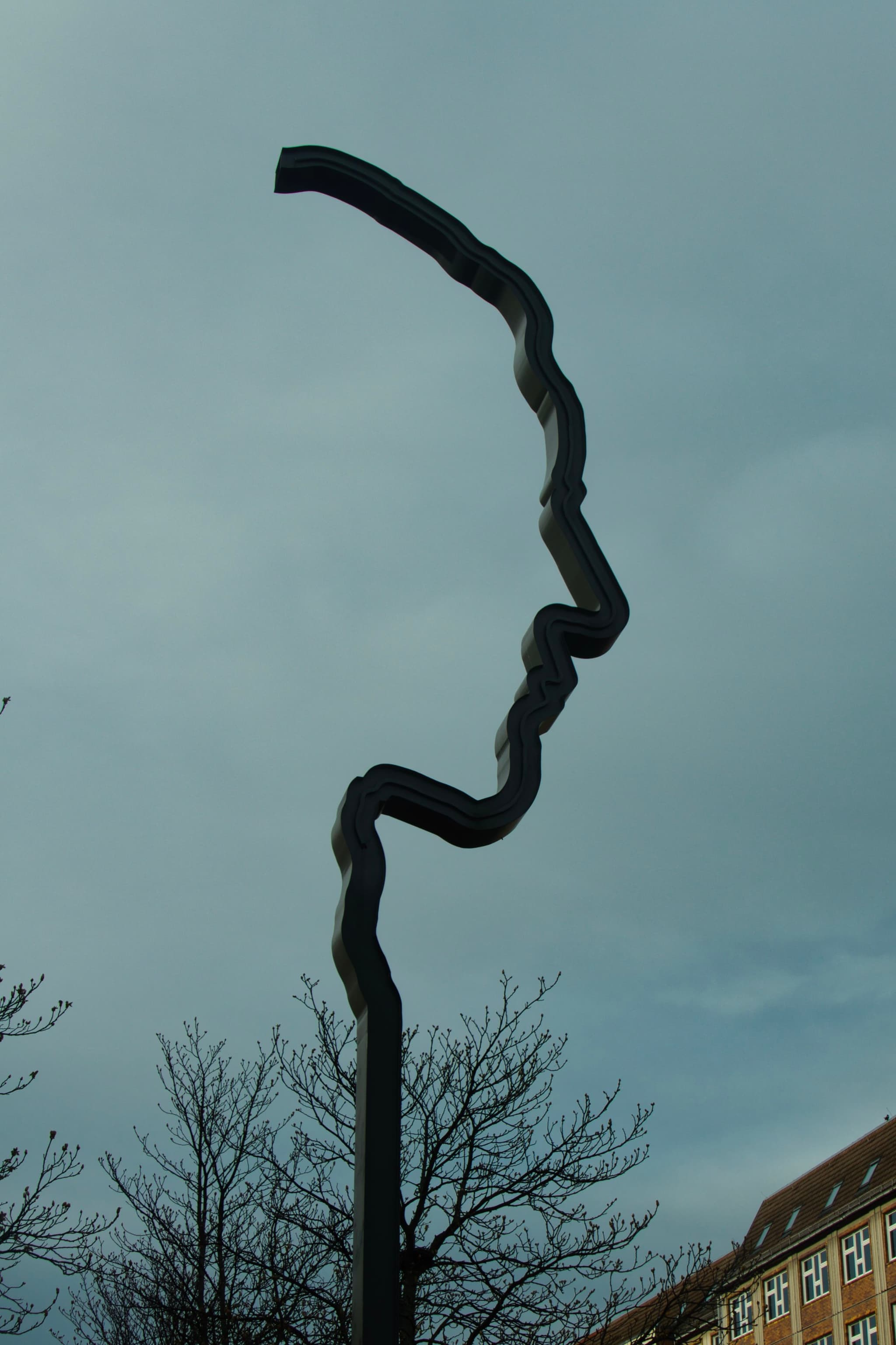 A metal sculpture shaped like a human profile against a cloudy sky, with bare trees and a building in the background