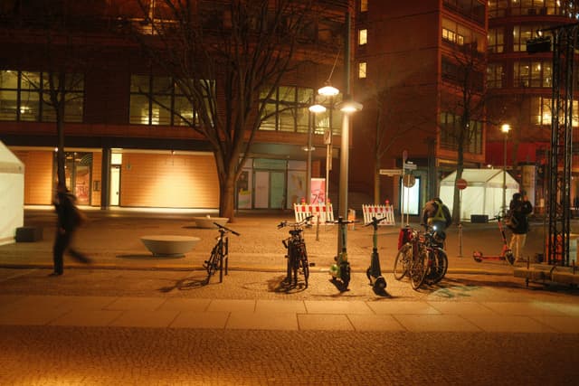 A nighttime urban scene with bicycles parked under streetlights, a person walking, and buildings in the background