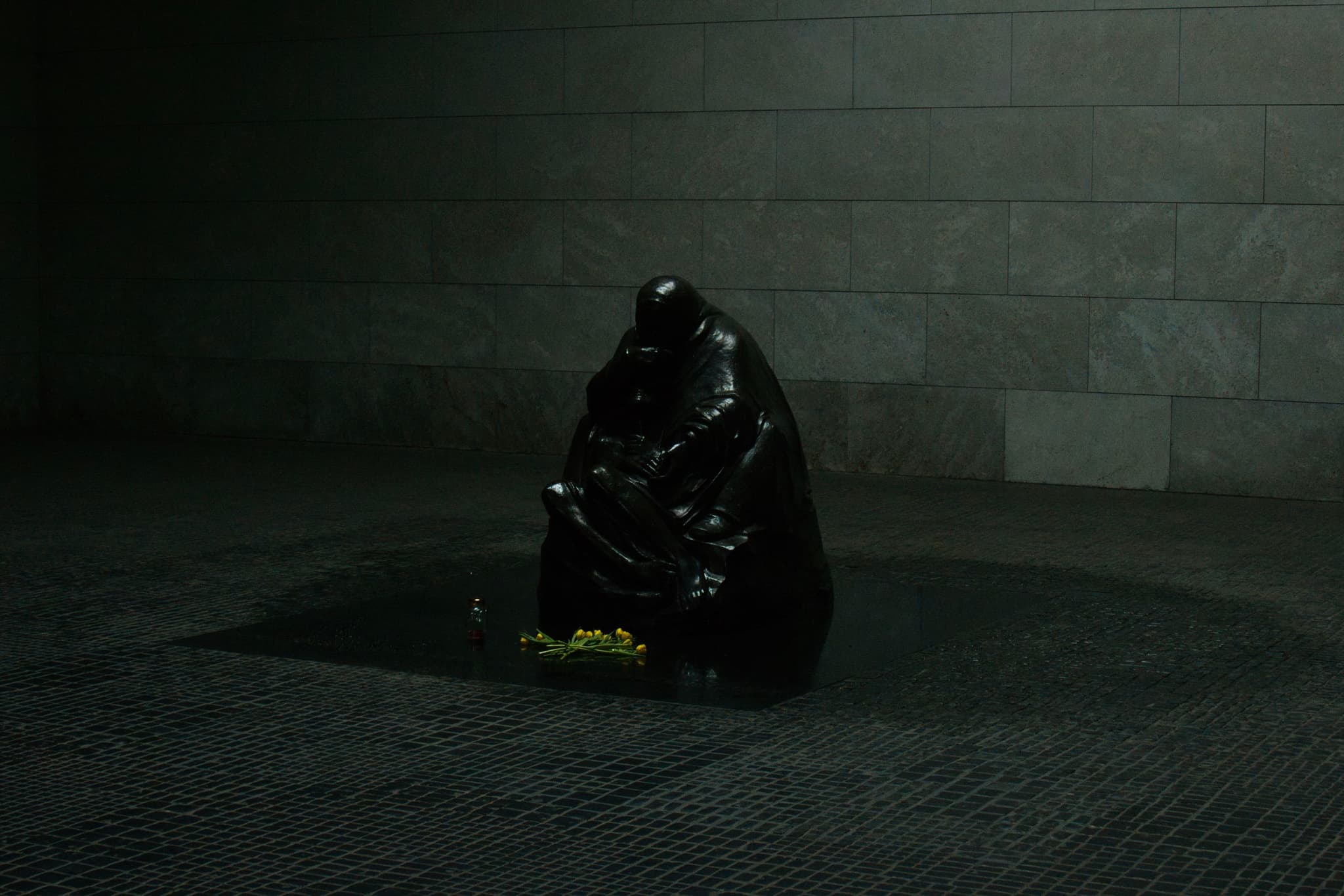 A dark, reflective sculpture of a seated figure in a dimly lit room with a small bouquet of yellow flowers at its base