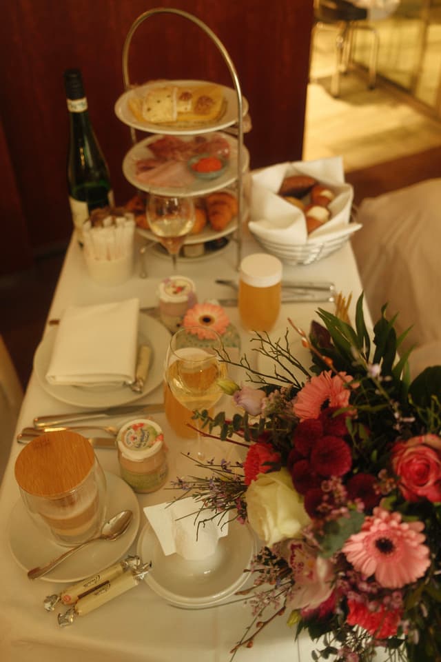 A table set for afternoon tea with a tiered stand of pastries, a bottle of wine, glasses, and a bouquet of flowers