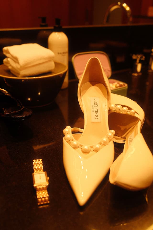 Elegant high-heeled shoes with pearl embellishments are placed on a glossy surface, accompanied by a gold watch. In the background, folded towels and a bottle are visible
