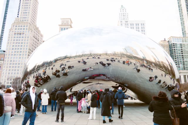 A large, reflective bean-shaped sculpture surrounded by people in an urban setting with tall buildings in the background