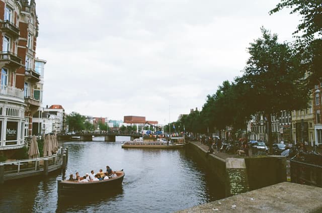 A canal scene with people in a boat, surrounded by buildings and trees, under a cloudy sky
